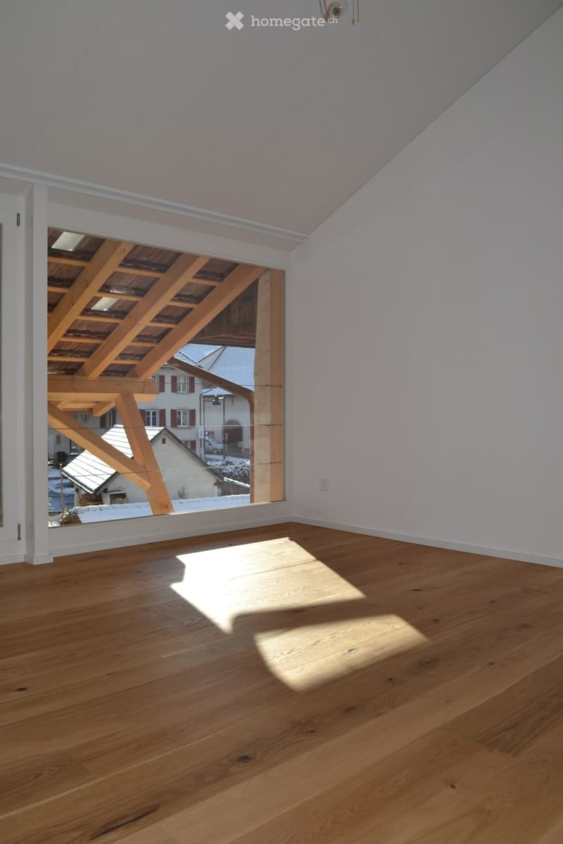 Empty room with wooden flooring and large window showing wooden beams and outdoor view.