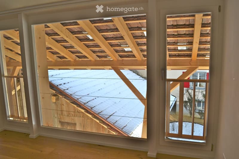 Empty room with wooden flooring and large windows offering views of a snowy roof and neighboring building.