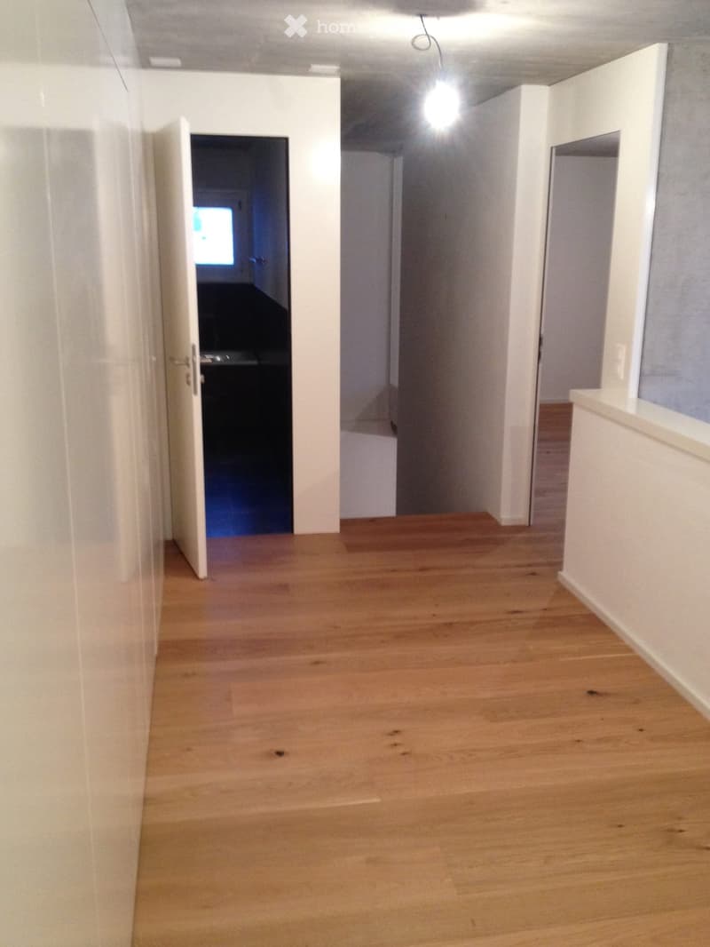 Empty hallway with light wooden flooring and white walls, featuring an open door leading to another room.