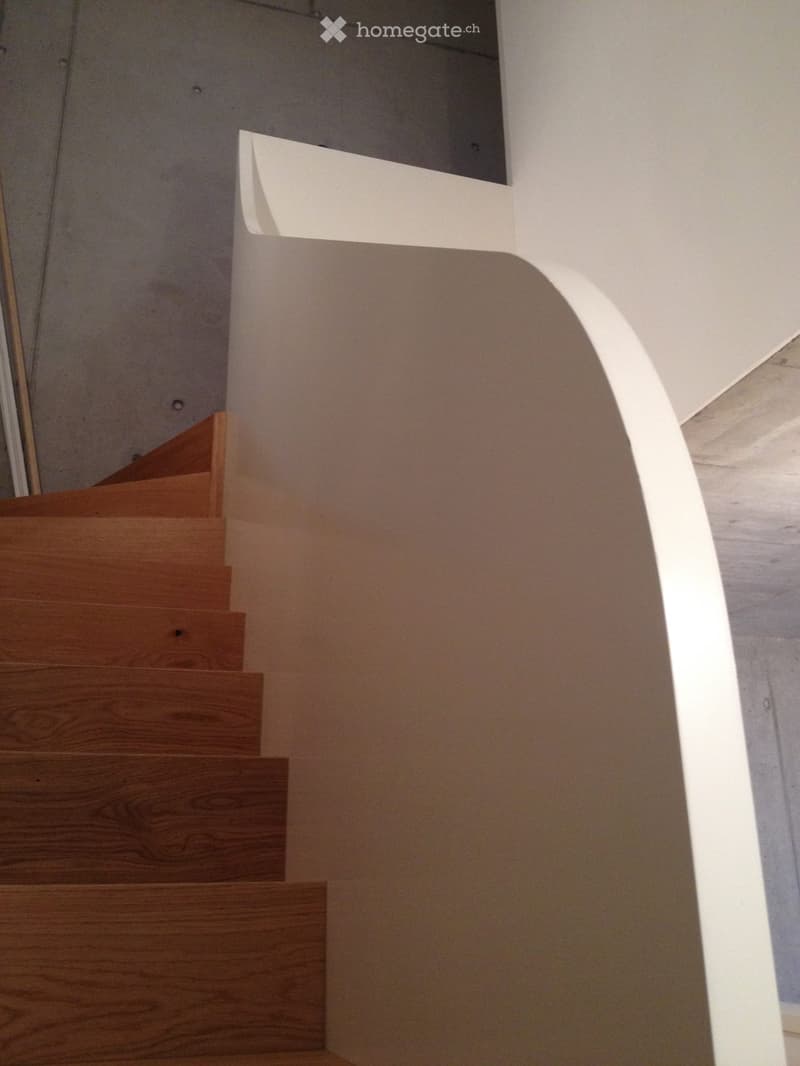 Modern staircase with wooden steps and a white curved handrail, leading upwards in a well-lit interior space.