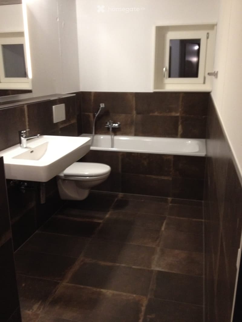 Modern bathroom with dark brown tiles, white sink, toilet, and bathtub, featuring a sleek faucet and a window with frosted glass.
