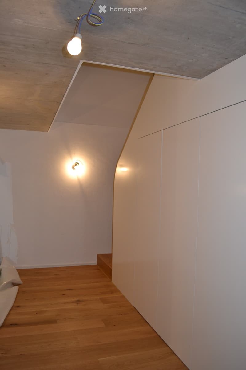 A narrow hallway with wooden flooring, white walls, and recessed lighting. The ceiling features exposed beams and a single hanging light fixture.