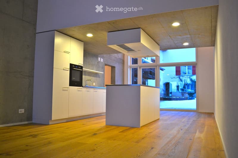 Modern kitchen with white cabinets, a built-in oven, a sink, and a central island, featuring wooden flooring and large windows.