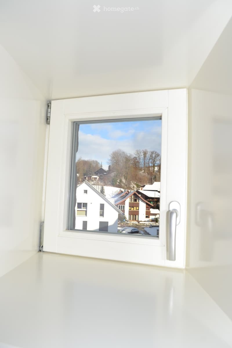 Empty room with white walls and a large window showing a view of houses and trees in the background.
