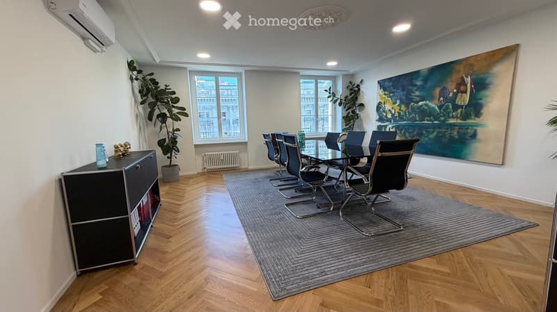 Modern furnished room with wooden flooring, large windows, a grey rug, a black cabinet, a large painting, and a set of black chairs around a glass table.
