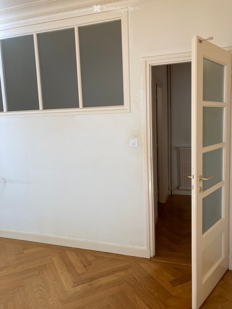 Empty hallway with wooden flooring, white walls, and a large window with frosted glass panels. An open door leads to another room with a radiator visible in the background.