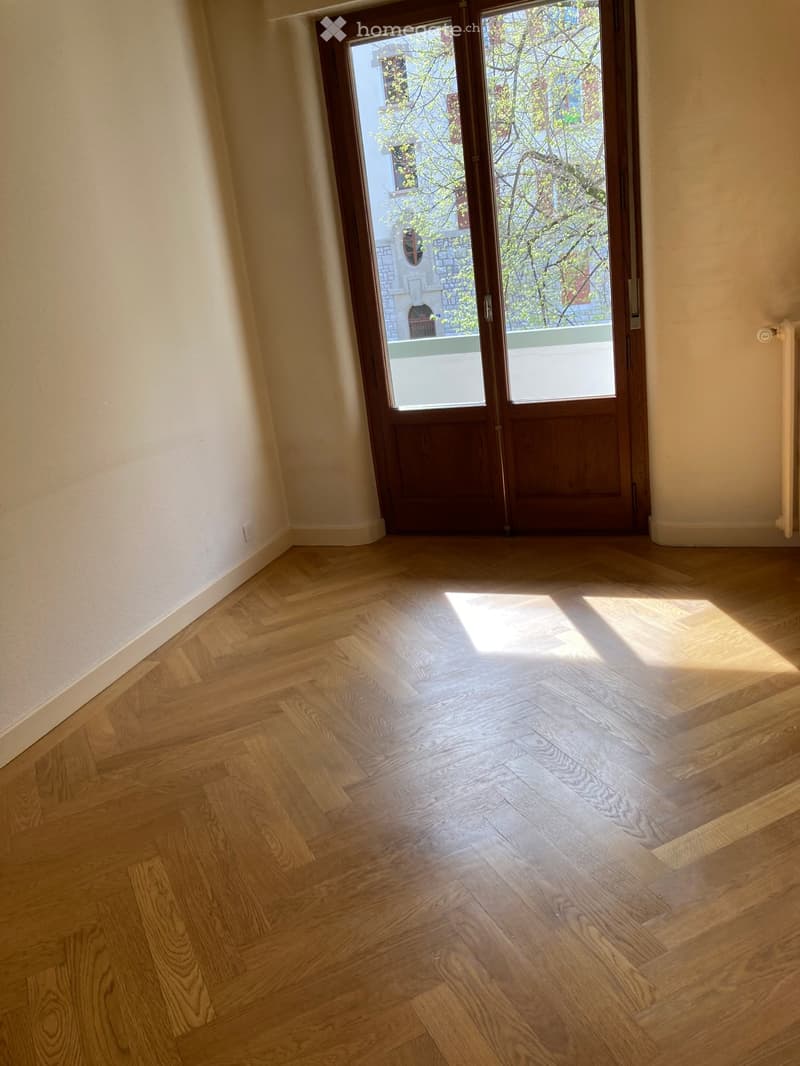 Empty room with light wooden herringbone flooring and a large glass door leading to a balcony or outdoor area. The walls are painted a light beige color, and the space is well-lit with natural light.