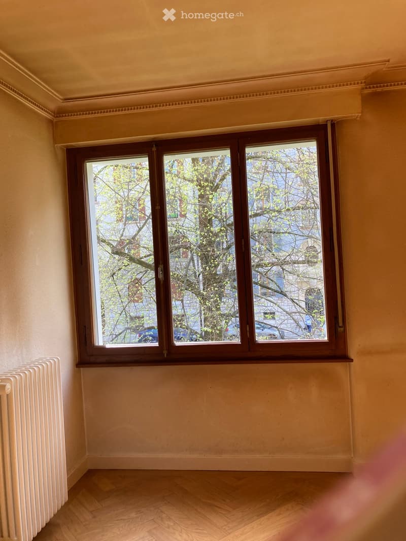 Empty room with wooden flooring, large window with wooden frame, radiator, and light-colored walls.