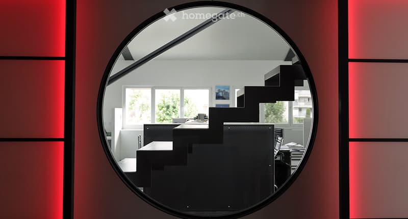 A modern, minimalist room visible through a round mirror, featuring a black chair, a table, and a screen, with large windows and artwork on the walls.