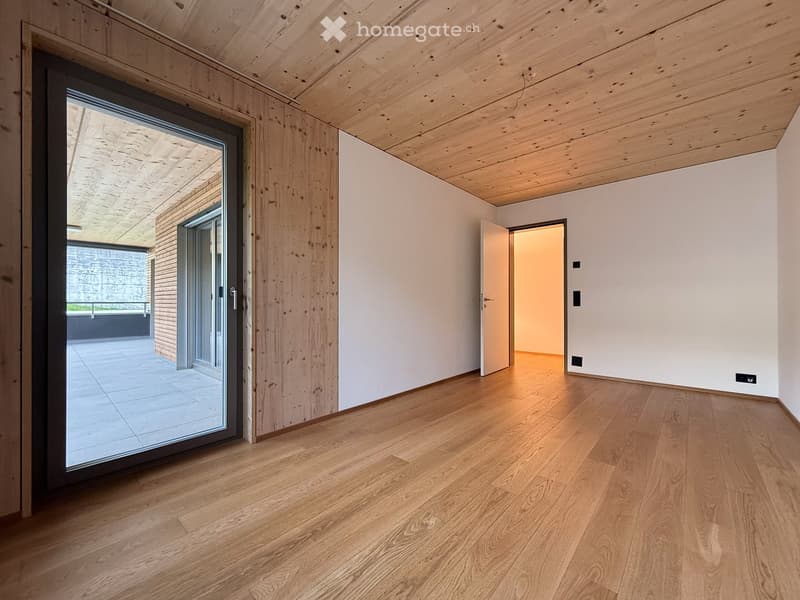 Empty hallway with light wooden flooring, white walls, and exposed wooden ceiling beams. Features a large sliding glass door leading to an outdoor area and an open doorway to another room.