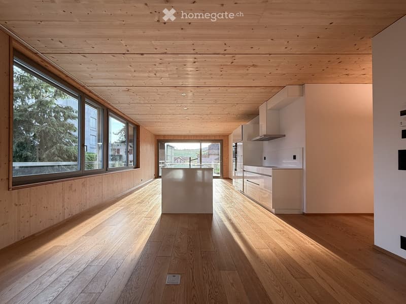 Open-plan kitchen and living area with light wood flooring, white kitchen cabinets, and a central island. Large windows and a sliding glass door provide natural light and access to the outdoors.