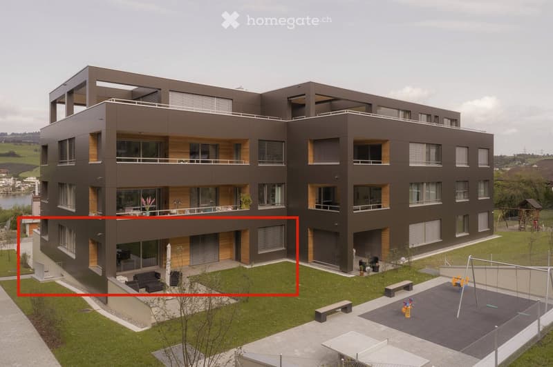 Modern multi-story residential building with balconies, large windows, and a landscaped courtyard featuring benches, a swing set, and a patio area.
