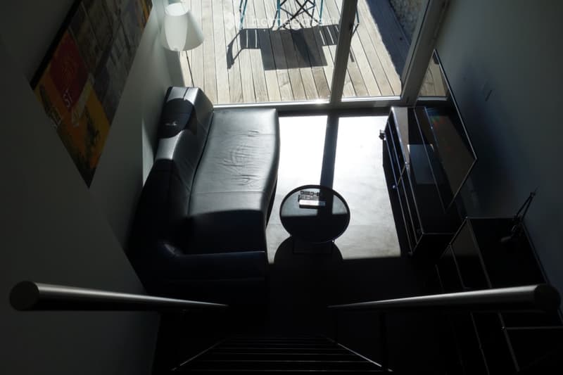 Modern living room with a black leather sofa, a round coffee table, a glass table, and a lamp near the wall. The room has large windows leading to a wooden deck with outdoor furniture.