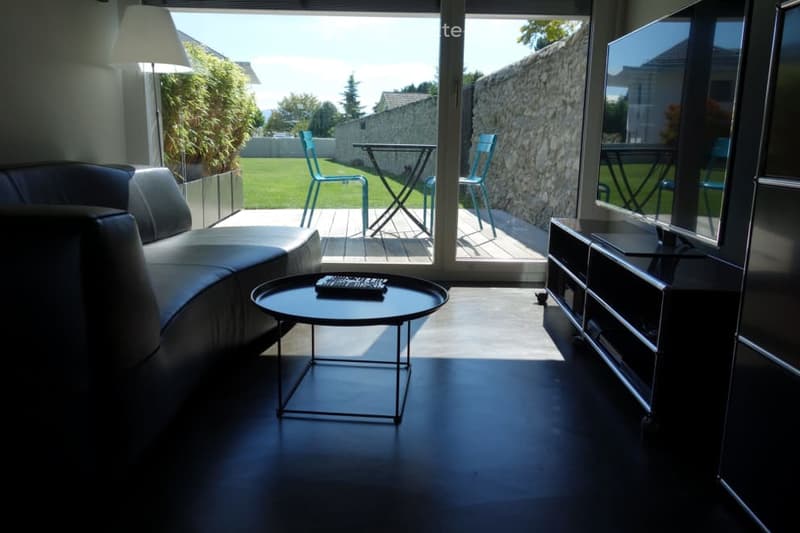 Modern living room with dark flooring, large glass doors opening to a patio, sofa, coffee table, TV stand, and outdoor furniture visible through the doors.