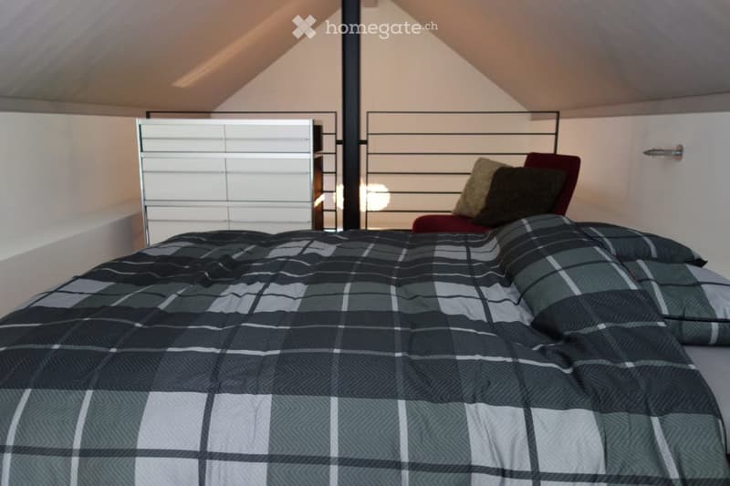 A modern bedroom with a large bed covered in a blue and white plaid duvet, multiple pillows, and a sleek chair with a red cushion. The room features white walls, a minimalist headboard, and a contemporary lamp.