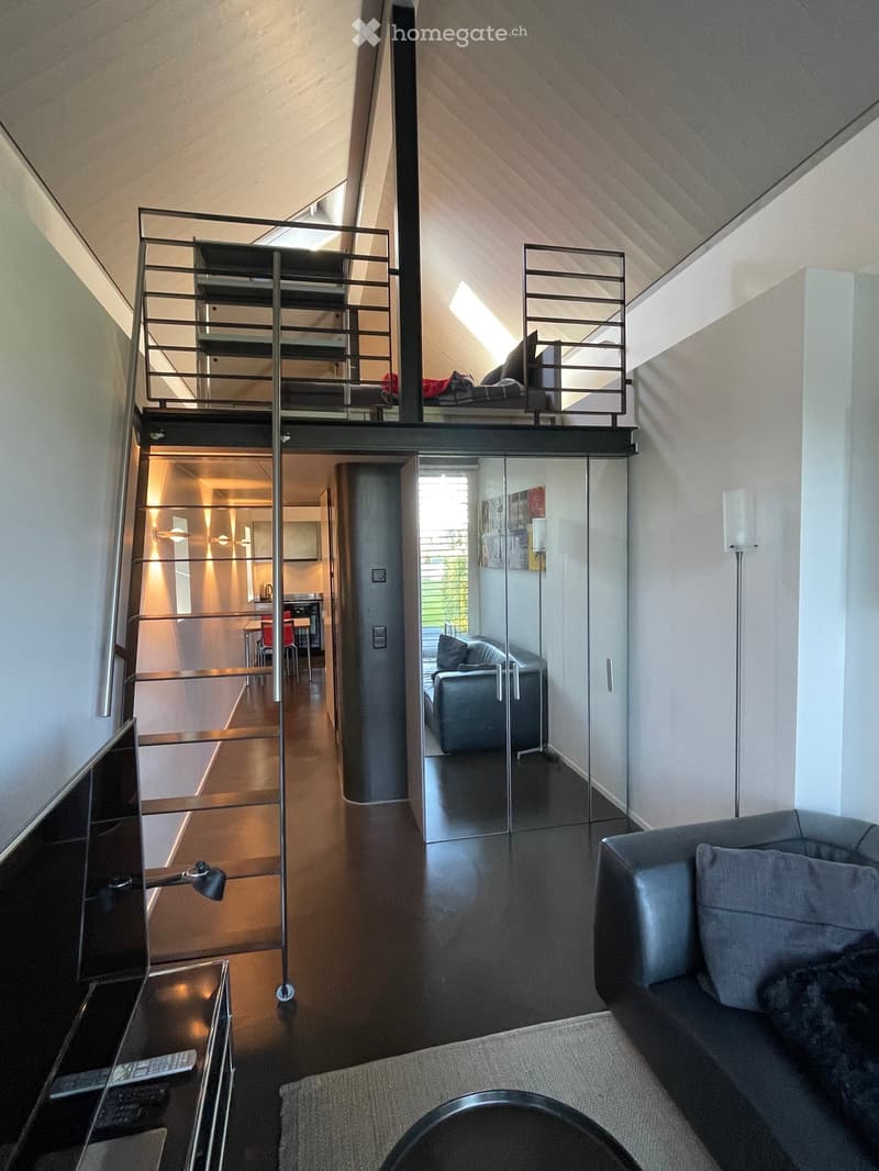 Modern living room with a sofa, coffee table, and a staircase leading to a lofted bedroom area. The space features sleek, contemporary furniture and ample natural light.