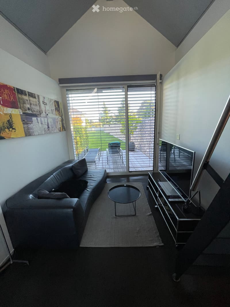 Modern living room with a black leather sofa, a round coffee table, and a TV stand, featuring large windows with blinds and a view of a garden.