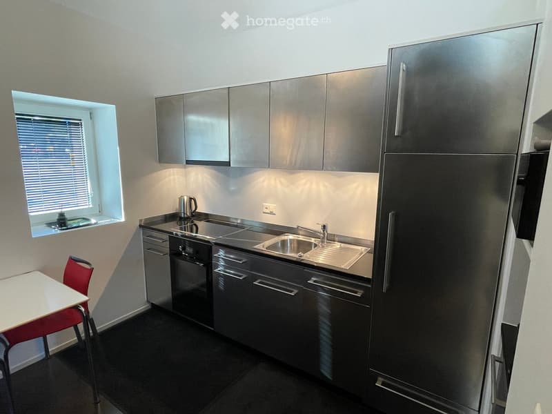 Modern kitchen with stainless steel appliances, including a refrigerator, oven, dishwasher, and sink. Features a small dining area with a table and chair, and a window with blinds.