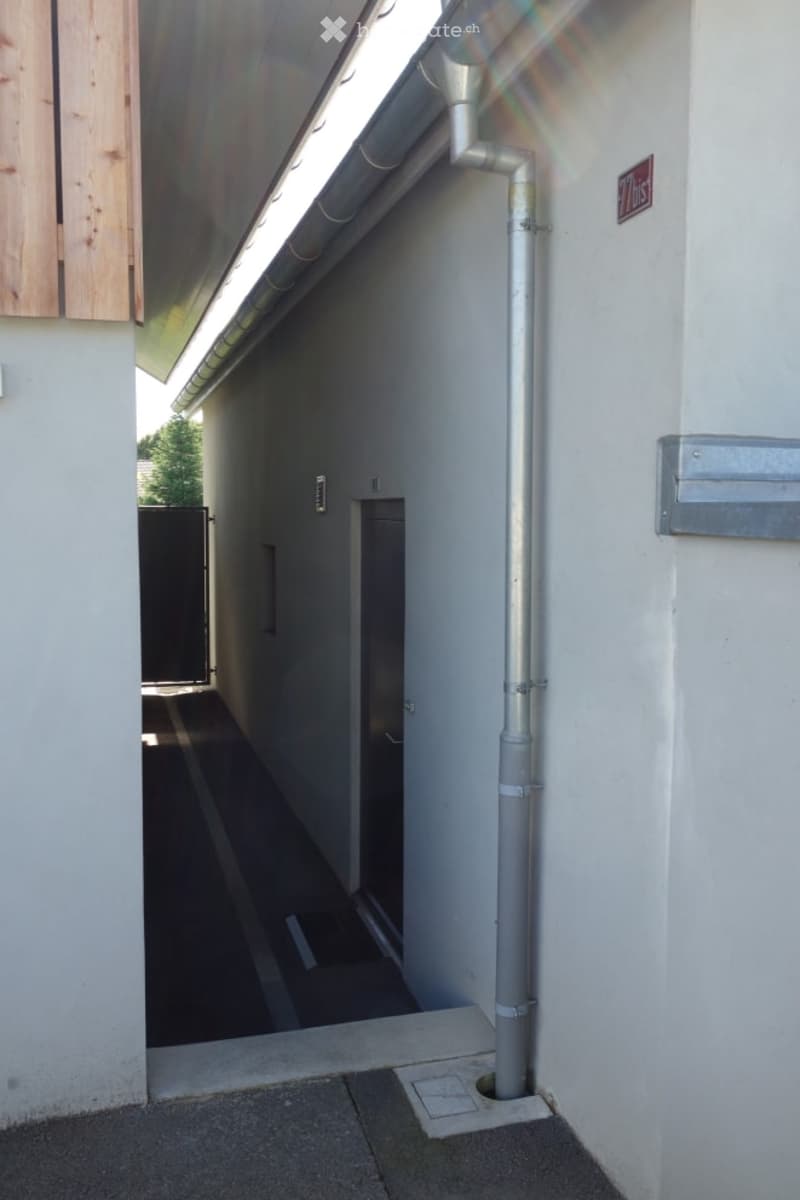 A narrow, covered outdoor corridor with white walls, a metal pipe on the right, and a small gate at the end.