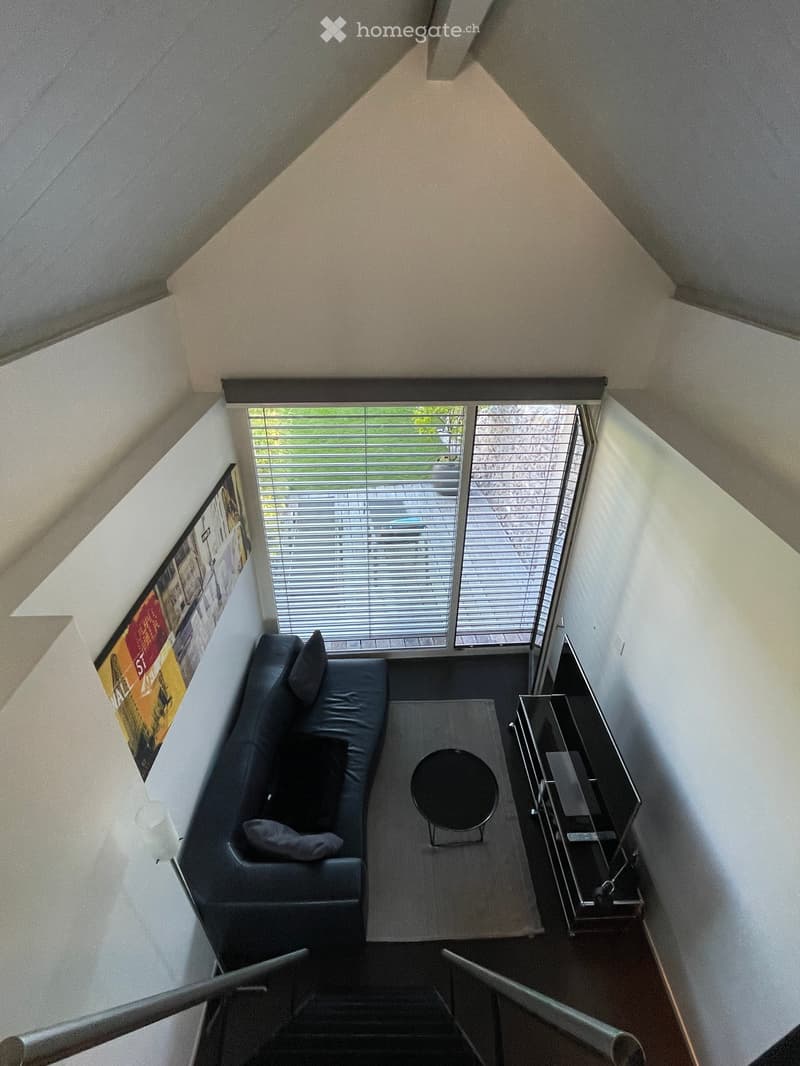 A modern living room with a black sectional sofa, a coffee table, a TV stand, and a small round table with a stool. The room has large windows with blinds and a view of the outside.