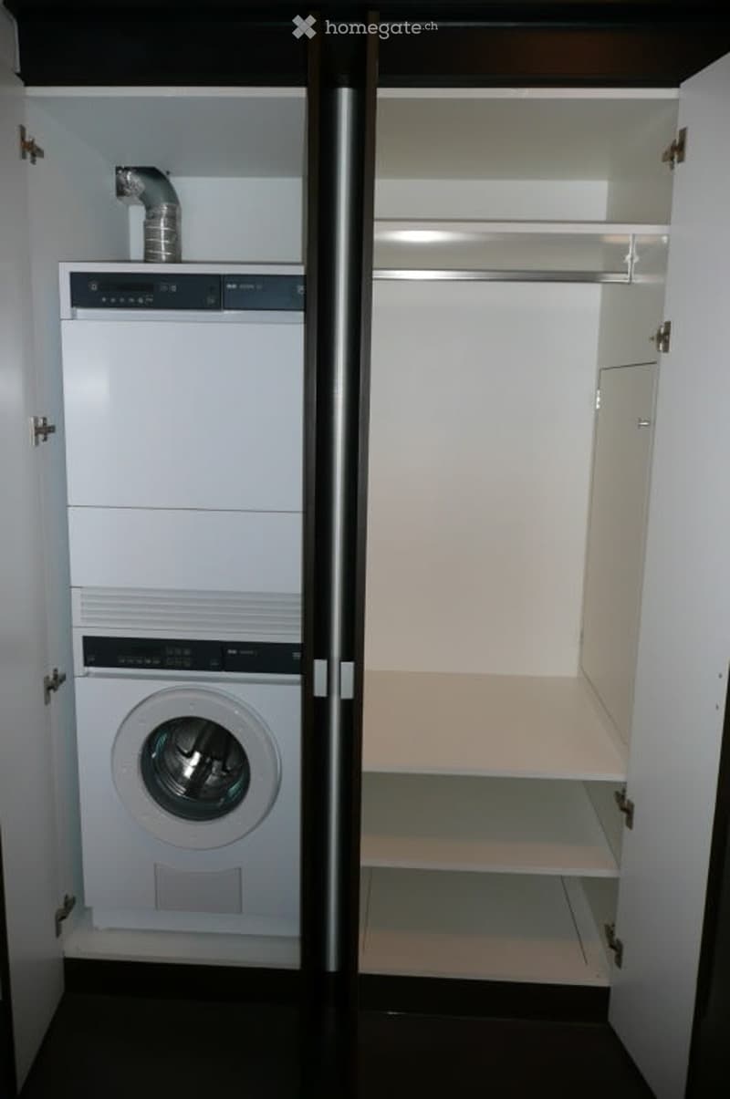 A compact laundry room with a white washing machine and dryer stacked vertically, a built-in closet with shelves, and a silver ventilation pipe on the wall.