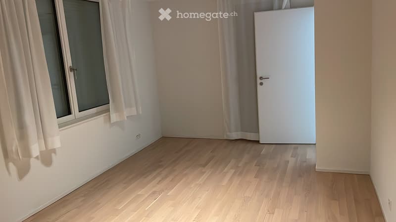 Empty room with wooden flooring, white walls, a window with white curtains, and a white door.