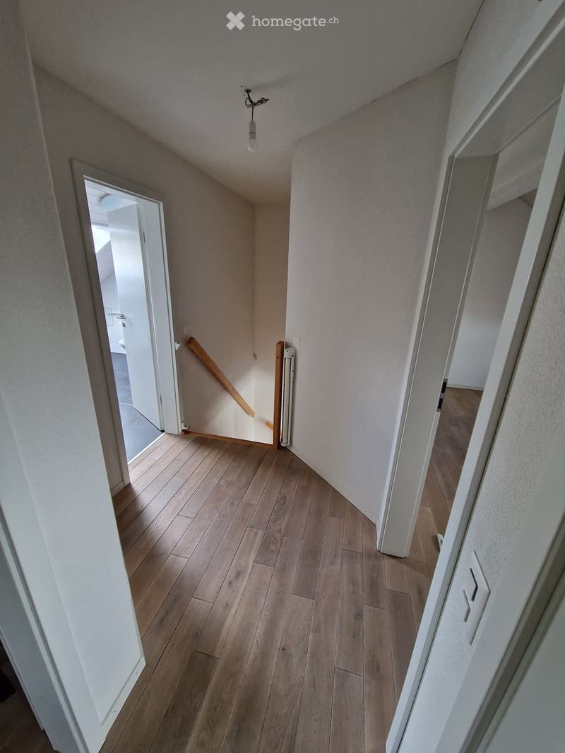 A narrow hallway with wooden flooring and white walls, featuring a single light fixture and an open doorway leading to another room.