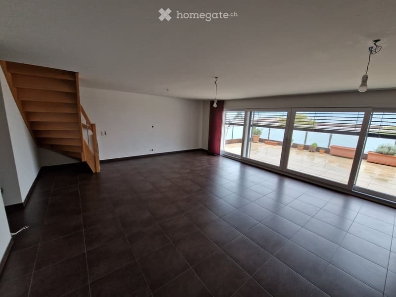 Empty room with dark tiled floor, wooden staircase, large sliding glass doors leading to a patio with outdoor plants.