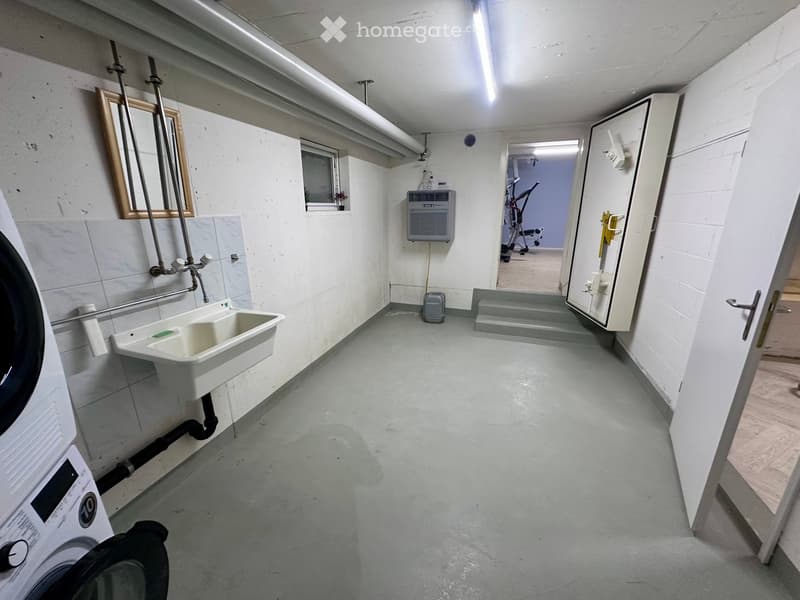 A small basement room with gray floor, white wall, sink, washing machine, mirror, pipes, air conditioner, window, and doors.