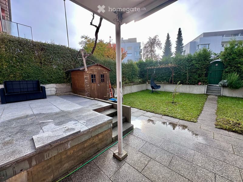 covered terrace with stone flooring, wooden storage shed, landscaped garden with hedges, lawn, and a swing