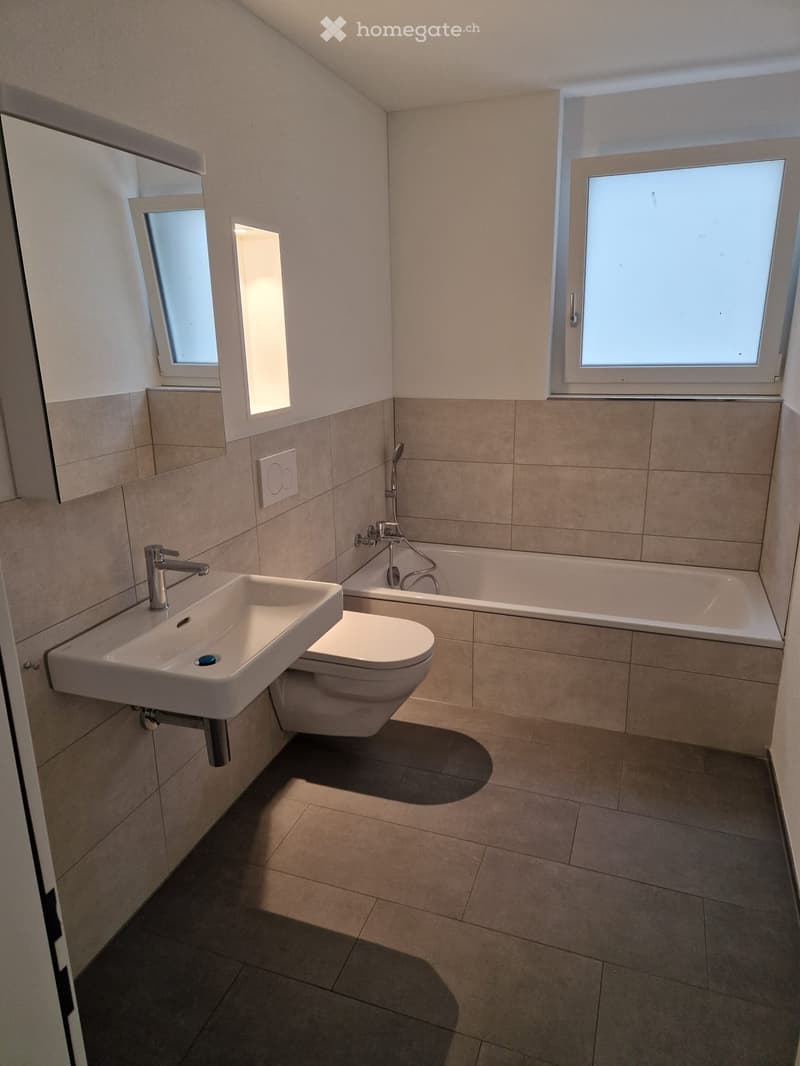 Modern bathroom with beige tiles, a white sink, toilet, bathtub, and a large mirror. Natural light from two windows illuminates the space.