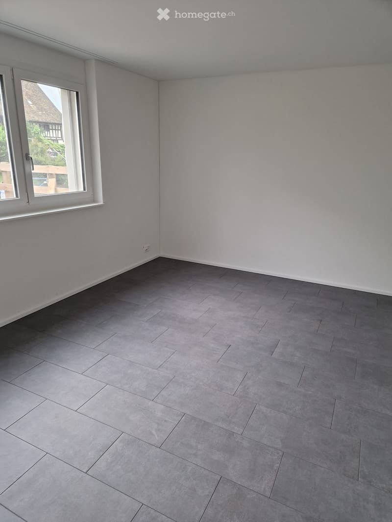 Empty room with grey tiled floor and white walls, featuring a large window with white frame and a visible electrical outlet on the wall.