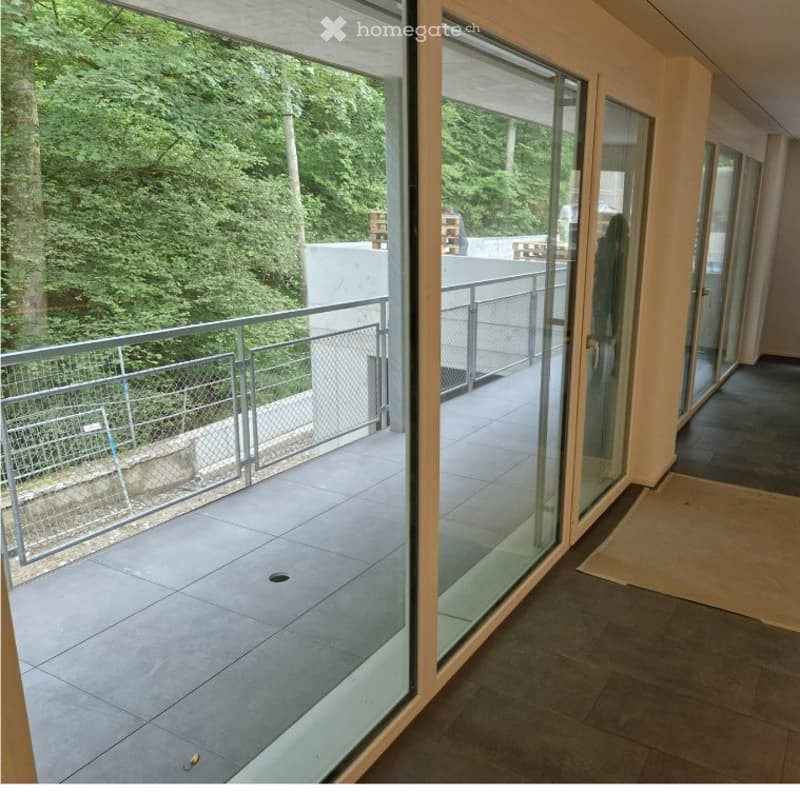 Modern balcony with large glass sliding doors, metal railing, and tiled flooring, overlooking a green, tree-filled area.