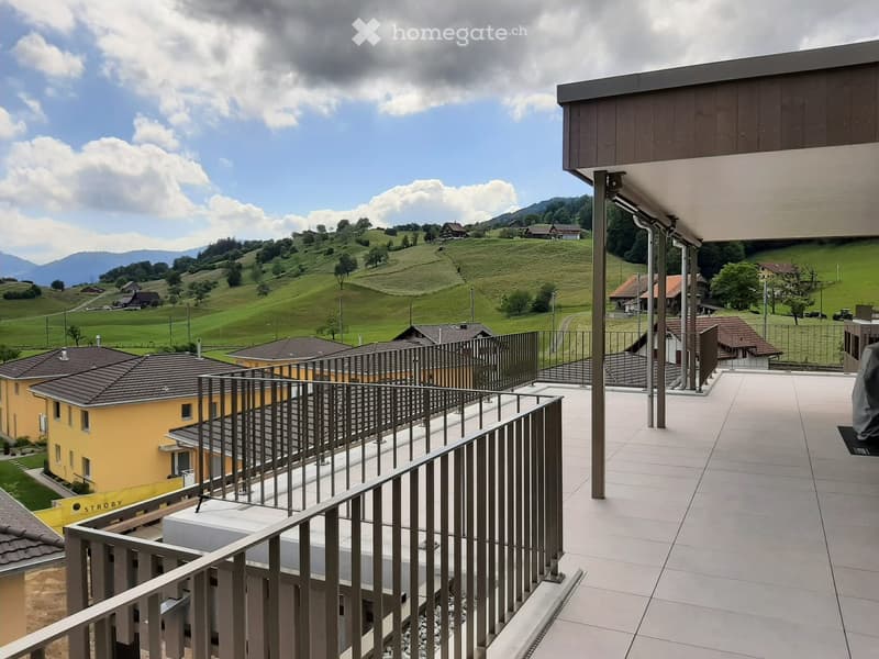 Balcony with modern metal railing, overlooking a scenic countryside with green hills, scattered houses, and distant mountains.