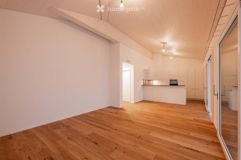 Open-plan unfurnished room with light wooden flooring, white walls, and a modern kitchen area featuring white cabinets and a countertop.