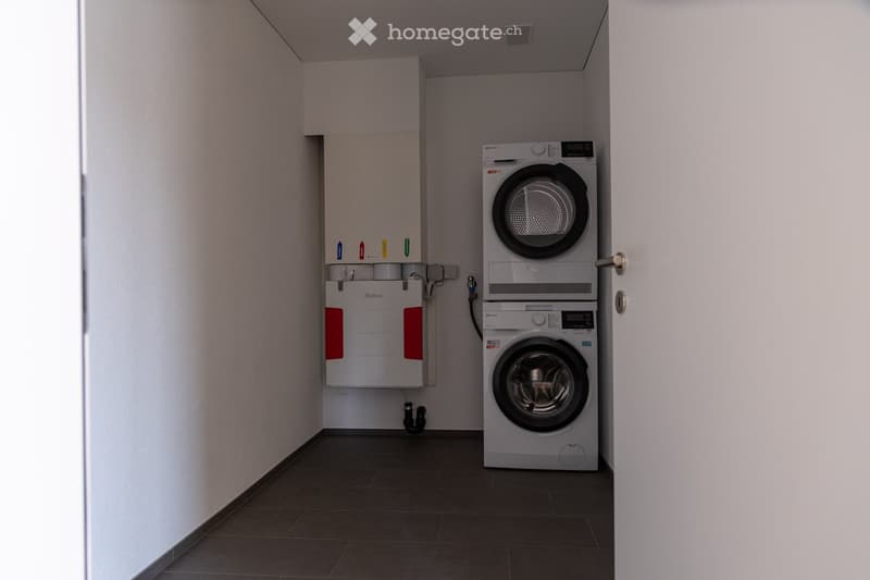A small, modern utility room with a washing machine, dryer, and water heater. The room has white walls, dark tiled flooring, and a door on the right side.