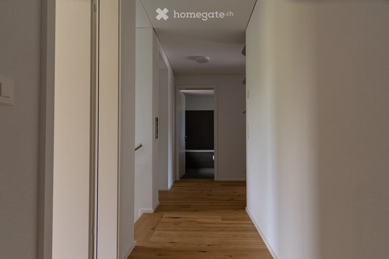 A clean, modern hallway with light wooden flooring, white walls, and recessed ceiling lighting. The hallway features several doors and a visible room at the end.