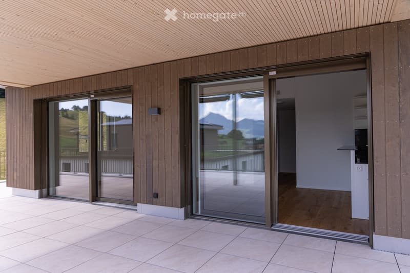 Modern balcony with large glass sliding doors, wooden panel walls, and tiled floor, offering a scenic mountain view.