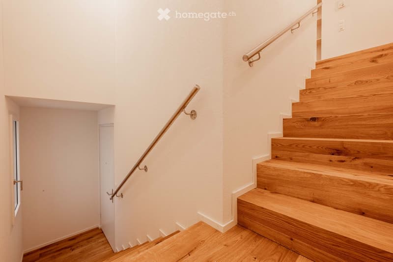 Modern wooden staircase with metal handrails, white walls, and wooden flooring. The staircase leads upwards with a visible landing and a door on the left side.