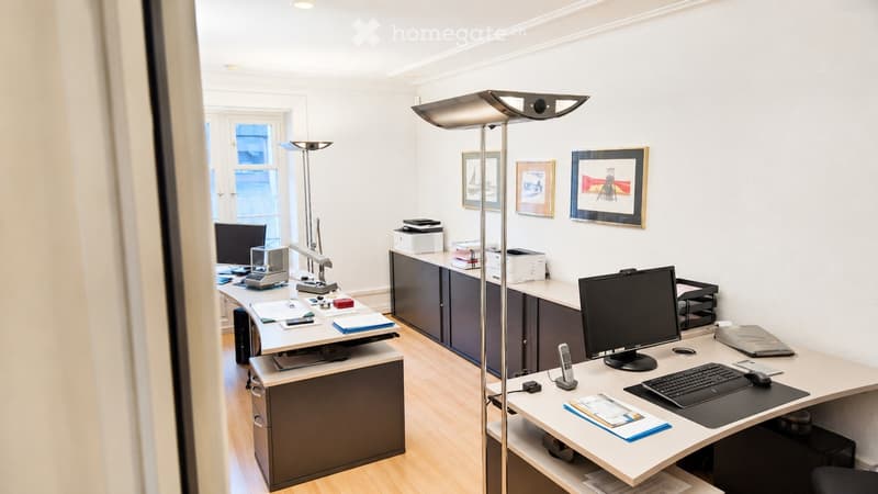 Modern office space with two desks, computer monitors, keyboards, mice, a floor lamp, cabinets, and framed artwork on the walls.