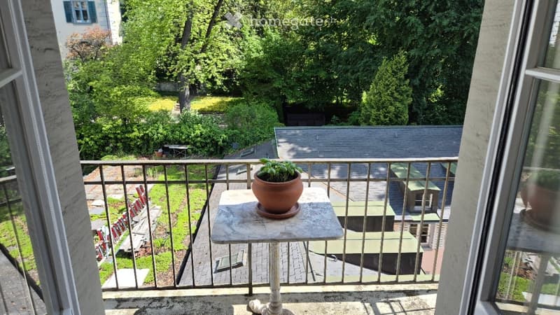 Balcony with a small table and potted plant, overlooking a green garden area with trees and a building in the background.