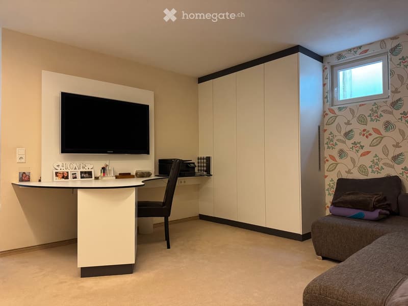 A modern, multi-functional room with a sofa, a desk with a chair, a television mounted on the wall, and a printer on the desk. The room features a floral-patterned wall and built-in cabinets.