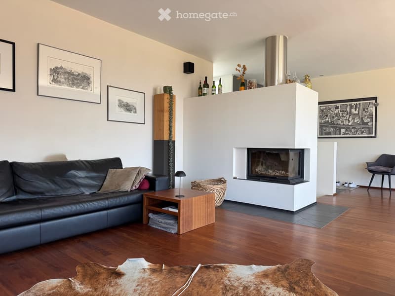 Modern living room with a black leather sofa, wooden coffee table, fireplace, and decorative items including framed pictures and wine bottles on the mantel.
