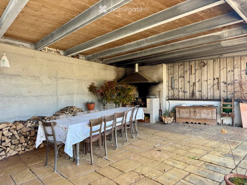 Outdoor dining area with a wooden table, chairs, a wooden cabinet, and a wood stack, featuring a rustic wooden ceiling and stone flooring.