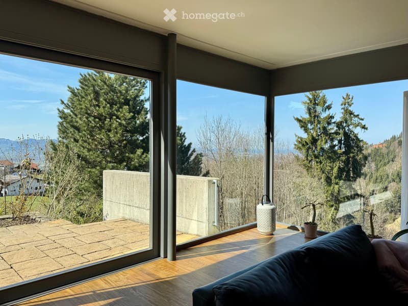 Bright living space with large windows offering a scenic view of trees and mountains, featuring a sofa, a small table with a teapot and potted plant, and wooden flooring.