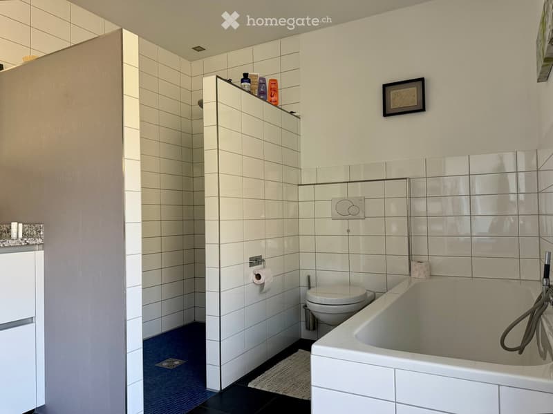 Modern bathroom with white tiles, a bathtub, toilet, and shower area. Includes a framed picture on the wall and a small shelf with toiletries.