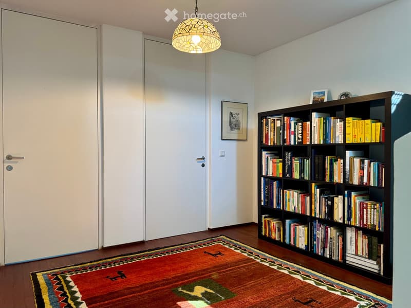A small, well-lit room with wooden flooring, a colorful patterned rug, a large bookshelf filled with books, a framed picture on the wall, and a decorative pendant light hanging from the ceiling.