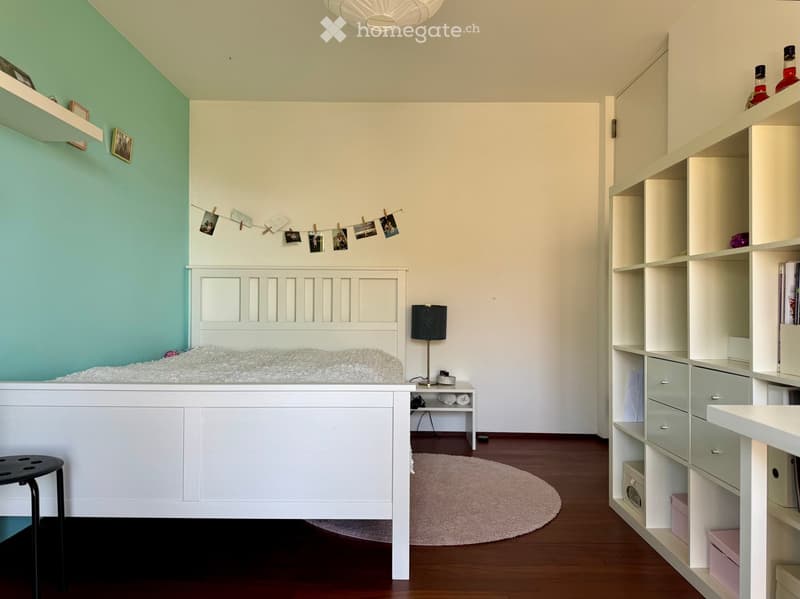 A modern, well-lit bedroom with a white bed, nightstand, lamp, and built-in shelving unit. The room features a light green accent wall, wooden flooring, and decorative elements like picture frames and a string of photos.