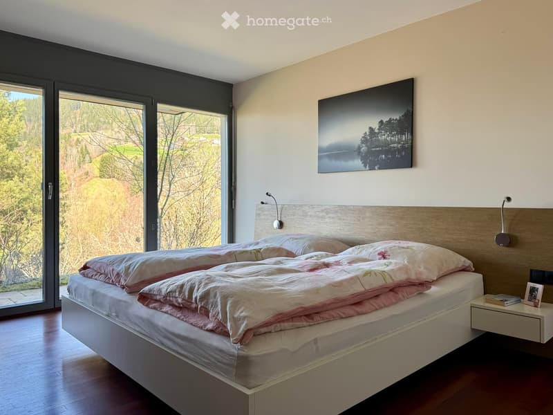 Modern bedroom with a large bed, pink bedding, a nightstand, and a large picture frame on the wall. Large windows offer a scenic view of the outdoors.