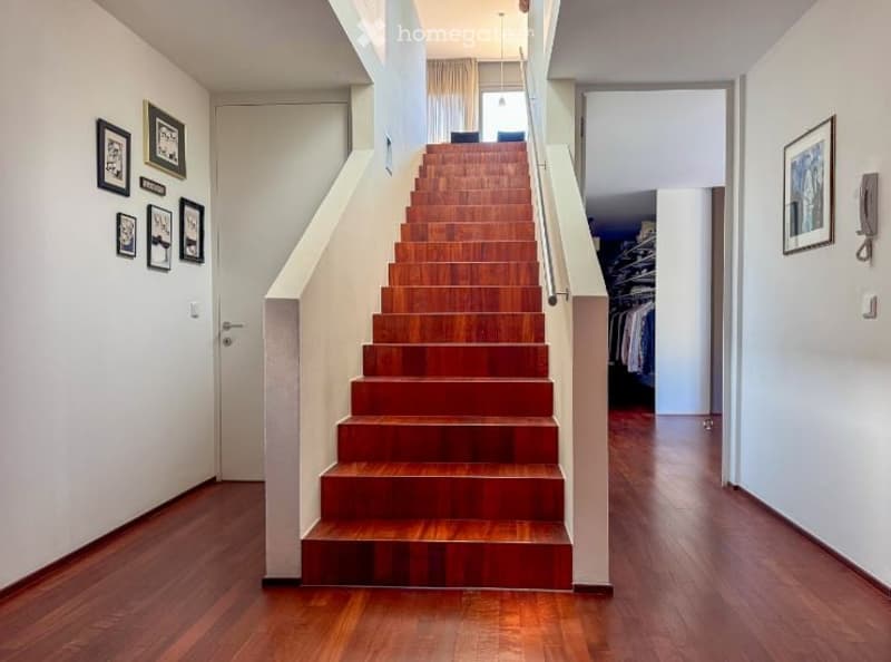 Spacious hallway with wooden flooring, a staircase leading upwards, and framed artwork on the walls. The staircase features wooden steps and a metal railing.